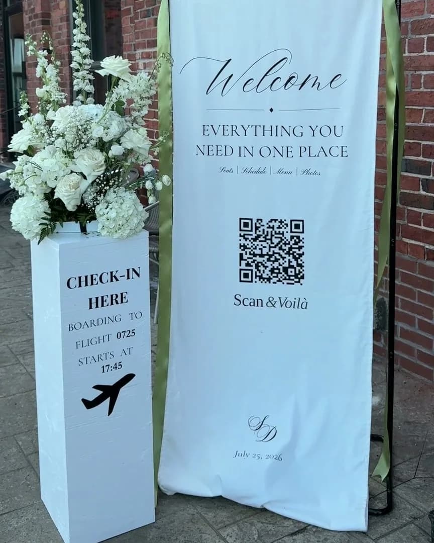 The Scan & Voilà sign at the door of our wedding on July 25, 2026: white flowers, a check-in stand and one QR code.